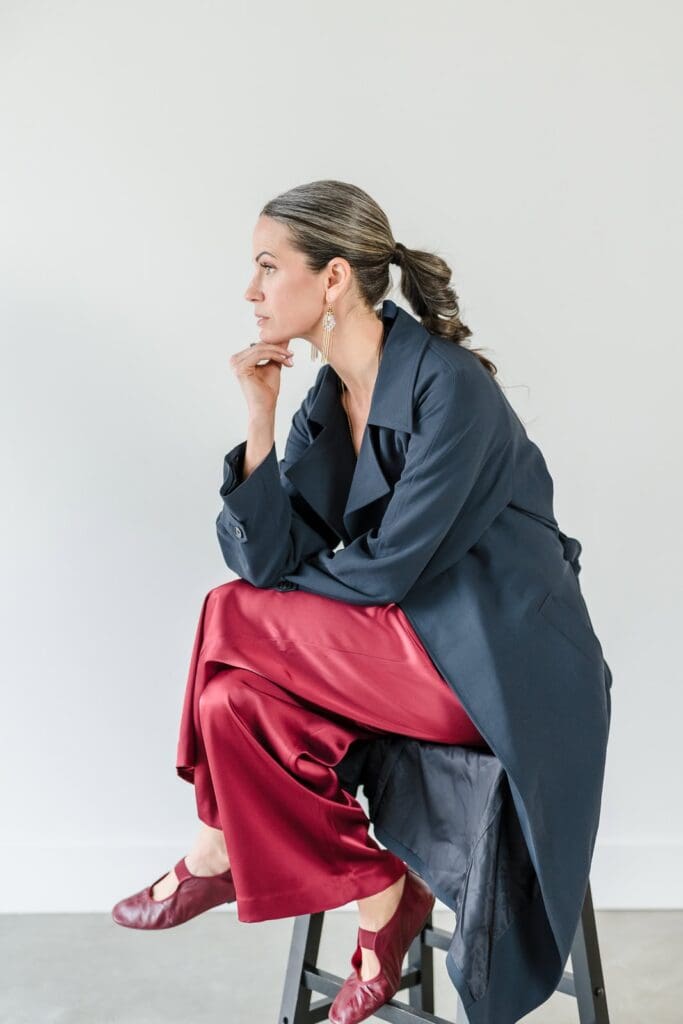 Confident woman sitting on stool side profile for brand photoshoot portrait in jewel tone silk outfit with matching shoes