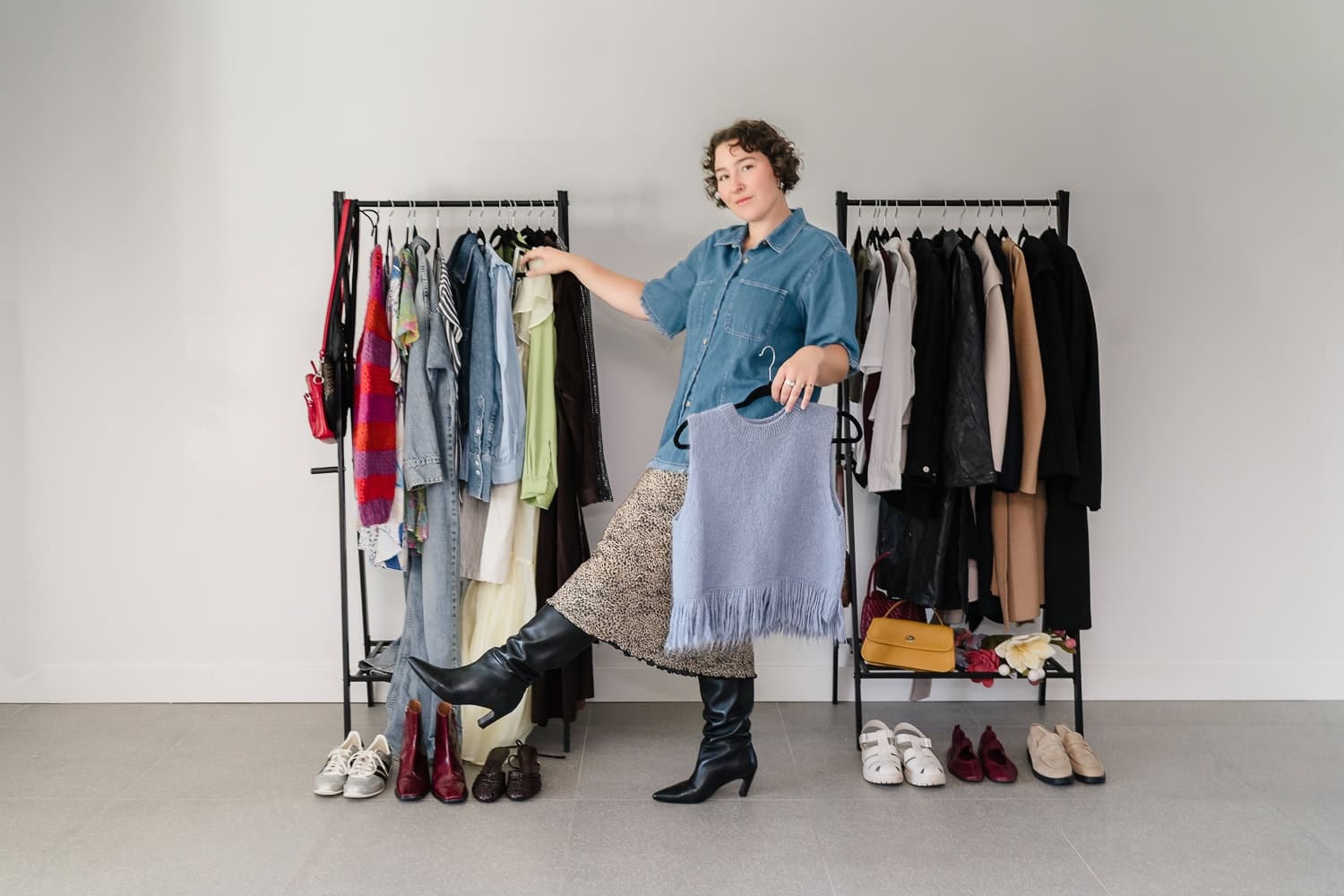 Woman stylist standing in front of racks of clothing and footwear holding a purple top and kicking her leg out.